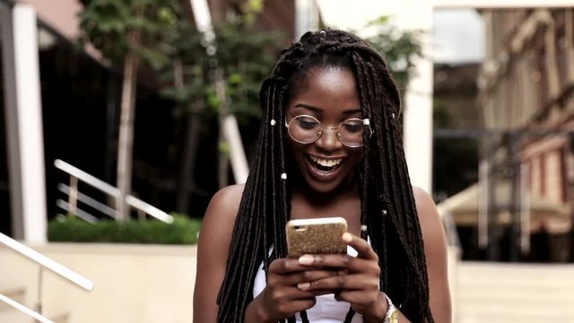 Portrait of Young Afro-american Girl Walking in the City. Happily Smiling Woman with Braids. Using her Smartphone. Wearing Round Eye Glasses.