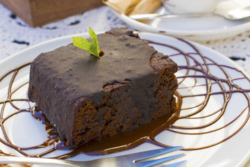 A piece of chocolate brownie on a white plate is decorated with a leaf of mint and caramel. Delicious chocolate dessert
