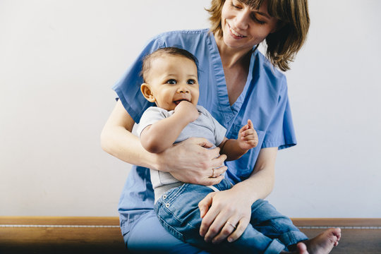 Smiling Female Doctor Holding Baby Boy While Sitting On Bench Against White Background