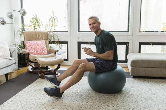Full length portrait of mature man sitting on fitness ball against windows at home