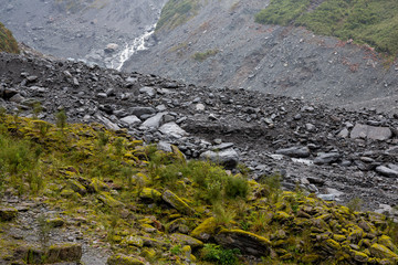 Rock formations with green foliage and stream at the base of Fox Glacier in New Zealand