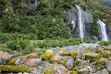 Waterfall and vegetation behind a slow moving stream at Franz Josef Glacier, south island New...