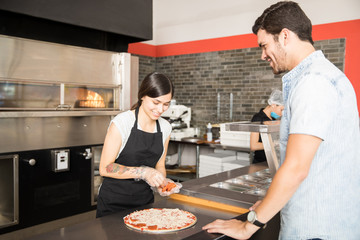 Smiling woman adding pepperoni slices to cheese pizza in kitchen counter