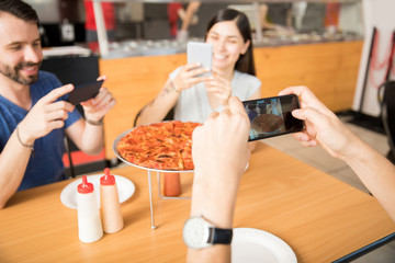 Hands of young man clicking pizza photo using smartphone with friends
