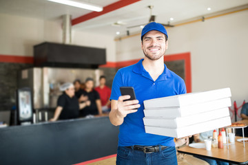 Smiling delivery man holding pizza boxes and smartphone looking at camera