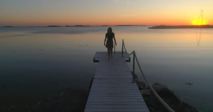 Girl on a wharf, C4K aerial descending tilt view a woman, walking on a wooden dock, enjoying the sun and a warm summer sunset, on a sunny evening dusk, in Hanko, Uusimaa, Finland