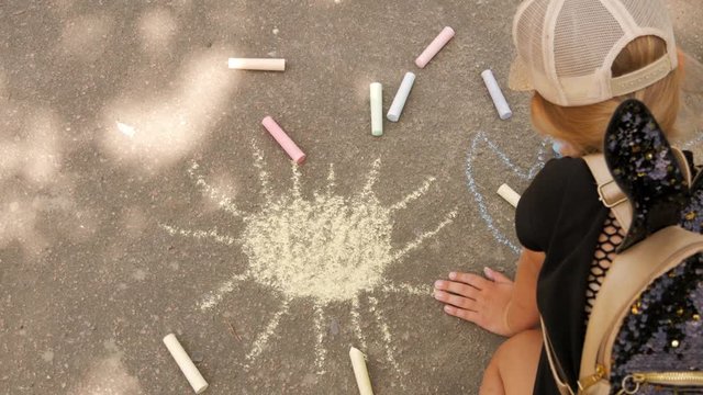 A little girl draws chalk on the asphalt. The child draws the sun in the park.