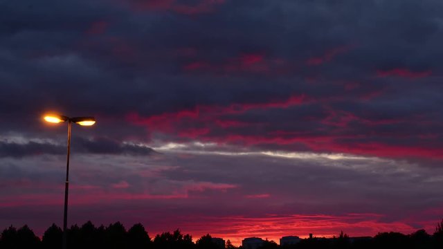 Sunset timelapse in summer, deep red sky and fast moving clouds with buildings in horizon and streep lamp in foreground.