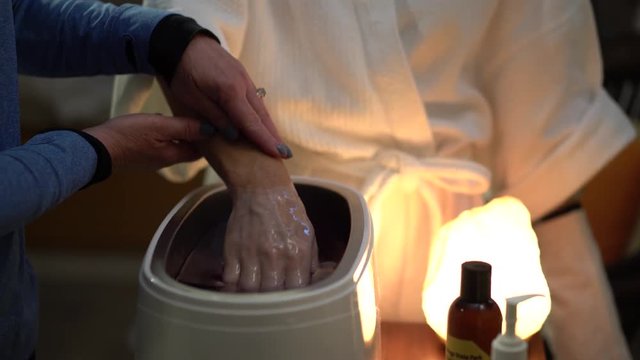 Camera sliding to the left of a closeup on a woman&rsquo;s hand as a therapist dips her hand into hot paraffin wax.
