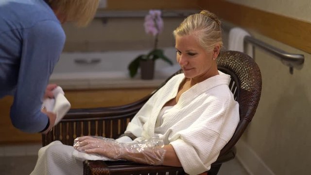 Pretty mature blonde woman sitting in a chair in a spa and a therapist taking gloves off of her hands after having a paraffin wax treatment on her hands.