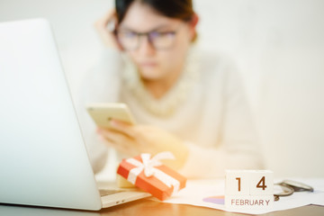 Business woman wearing glasses still working in Valentine day. Red gift box white ribbon using notebook laptop ,smartphone on office desk and wooden calendar 14 February. Hard work, holiday festival.