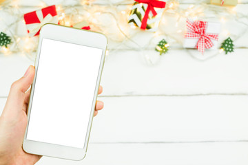 Close up hand holding mobile smartphone white screen with copy space for text Christmas New year theme red craft gift box ribbon bow with sparkling light on wooden table background.Displaying picture.