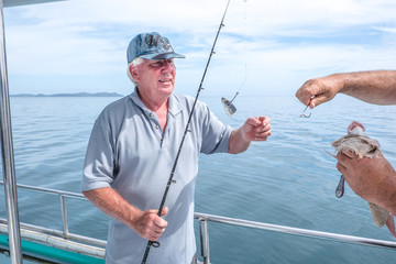 Fisherman helping caucasian tourist on fishing charter boat taking hook out of snapper fish - at Doubtless Bay, Far North, Northland, New Zealand, NZ