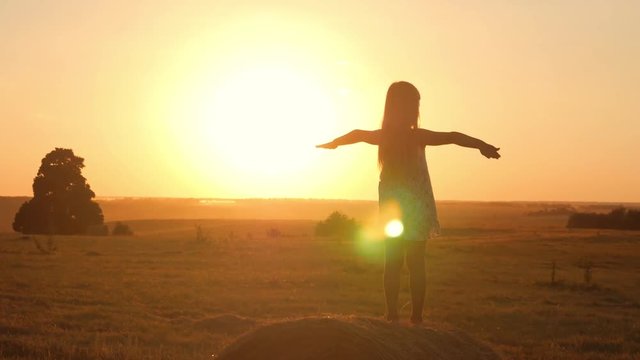 Young girl in a golden field standing on a reel of straw during sunset raising her hands in happiness. Beauty happy joyful little girl silhouette over sunset sky enjoying nature outdoors.