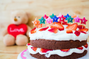 Homemade chocolate whipping cream with strawberry fruit cake decorate colorful candle with cute teddy bear and red love heart on wooden table background ,Happy Birthday party romantic creative design.