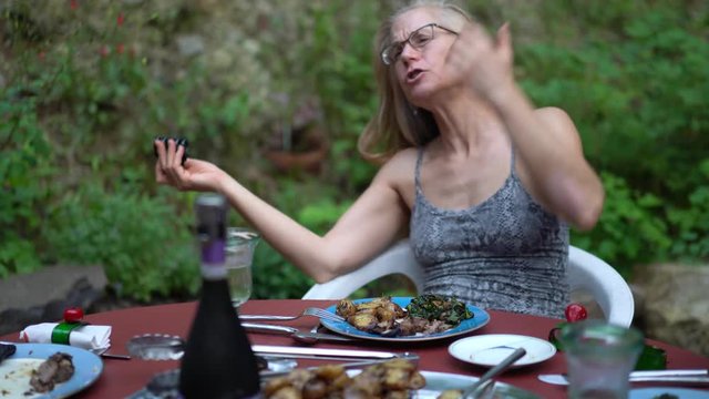 Mature blonde woman plays with her hair while eating and talking at a dinner table set outside in a garden.