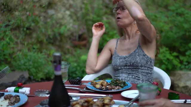Mature blonde woman plays with her hair while eating and talking at a dinner table set outside in a garden.