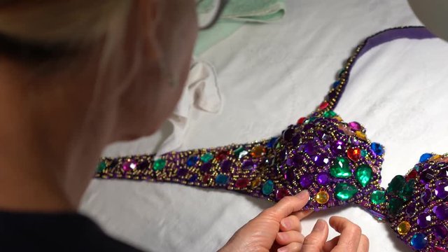 Looking down onto a decorated bra, a woman uses a glue gun to glue plastic rhinestones into place.