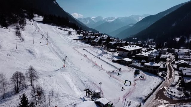 Drone orbit flying around small ski slope next to a small village in a beautiful valley, mountains in background. Clear sunny weather.