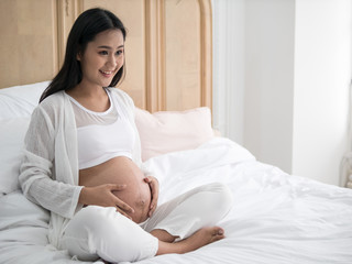 Happy pregnant asian woman sitting on bed and touching her belly in bedroom at home
