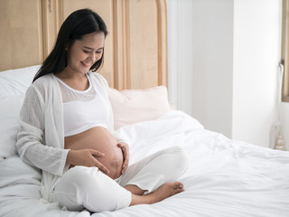 Happy pregnant asian woman sitting on bed and touching her belly in bedroom at home