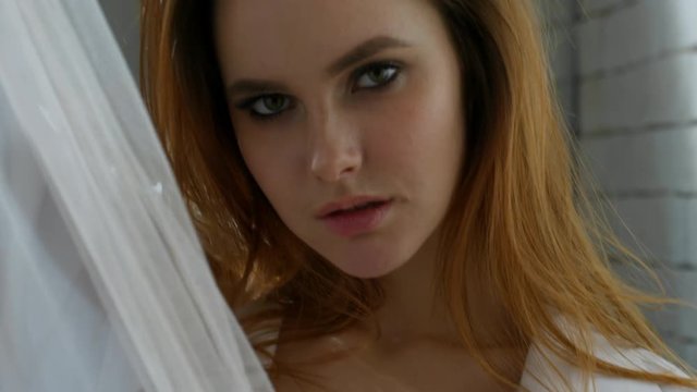 Tilt up with close up of sensual young woman in lingerie and shirt hiding behind sheer curtain and biting her lip while posing for camera