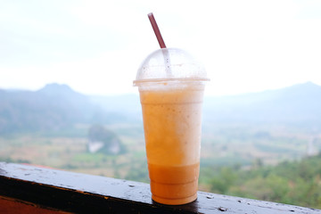 A cup of iced latte coffee on the fence of terrace in nature with mountain and flowers field background.