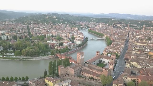 Aerial shot of Verona, Italy at sunset with typical Italian apartment rooftops, street crowds, Roman Colosseum Arena, Adige River, Santuario Madonna di Lourdes, mountains Lessinia Natural Park.