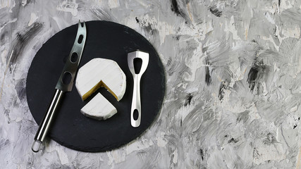 Piece of cheese camembert or brie in paper on a round shale board tasting with knives for cheese....