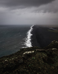 Bird perspective on a coastline in Iceland with a black sand beach with black and white contrasts between the waves and the beach while a storm is approaching with dark clouds in the sky
