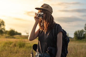 Young female tourist taking photo at hiking trip. Woman with backpack stands at sunset and photographs beautiful rural area