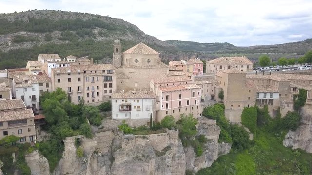 Aerial view of Cuenca's historical cathedral which in on a cliff in the middle of a valley in Spain. Show are colorful pastel houses and other buildings.