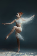 Dancing in flour concept. Redhead cute female girl adult woman dancer in dust / fog. Girl wearing white top and shorts making dance element in flour cloud on isolated grey background
