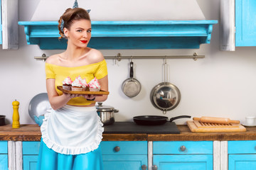 Retro pin up girl woman female housewife wearing colorful top, skirt and white apron holding tray with cooked sweet cupcakes standing in the kitchen with blue cabinets and utensils.