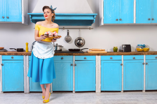 Retro pin up girl woman female housewife wearing colorful top, skirt and white apron and yellow high heels holding tray with sweet cupcakes standing in the kitchen with blue cabinets and utensils.