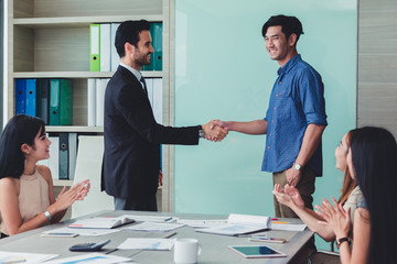 Great job two cheerful businessmen shaking hand while their colleagues applauding smiling..