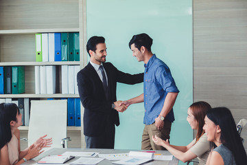 Great job two cheerful businessmen shaking hand while their colleagues applauding smiling..