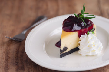 Delicious and sweet blueberry New York cheesecake on white plate served with whipped cream on wood table in close up view with copy space. Homemade bakery for cafe and restaurant or birthday cake.