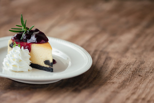 Delicious and sweet blueberry New York cheesecake on white plate served with whipped cream on wood table in side view with copy space. Homemade bakery for cafe and restaurant or birthday cake.
