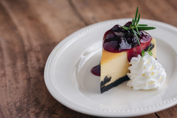 Delicious and sweet blueberry New York cheesecake on white plate served with whipped cream on wood table in close up view with copy space. Homemade bakery for cafe and restaurant or birthday cake.