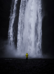 Famous Skogafoss Waterfall in Iceland with a small man in a yellow coat looking at the water falling off the hill in a dark look with black stones in the foreground and no people in front during night