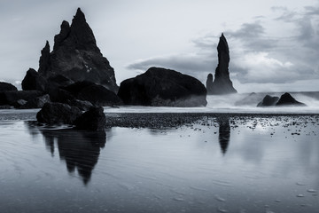 Rocks on a black sand beach in Iceland with reflection in the Sea and a dark sky in misty moody weather with dark colors and a rough sea in landscape format with a storm approaching