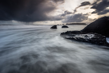 Rocks on a black sand beach in Iceland with reflection in the Sea and a dark sky in misty moody weather with dark colors and a rough sea in landscape format with a storm approaching