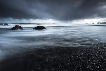 Rocks on a black sand beach in Iceland with reflection in the Sea and a dark sky in misty moody weather with dark colors and a rough sea in landscape format with a storm approaching