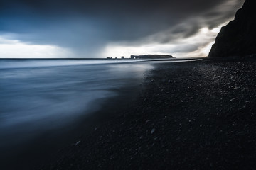 Rocks on a black sand beach in Iceland with reflection in the Sea and a dark sky in misty moody weather with dark colors and a rough sea in landscape format with a storm approaching
