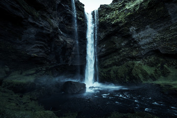 A wild natural fresh sparkling waterfall in a lonely green valley with moos covered rocks and ice cold mountain water flowing down black rocky walls on a rainy moody day in Iceland without tourists