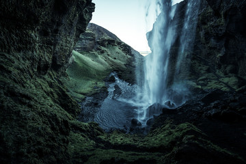 A wild natural fresh sparkling waterfall in a lonely green valley with moos covered rocks and ice cold mountain water flowing down black rocky walls on a rainy moody day in Iceland without tourists