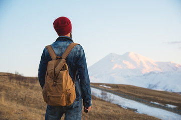 Bearded tourist hipster man in a hat with a backpack stand back on a roadside bump and watching the sunset against the background of a snow capped mountain