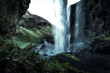 A wild natural fresh sparkling waterfall in a lonely green valley with moos covered rocks and ice cold mountain water flowing down black rocky walls on a rainy moody day in Iceland without tourists