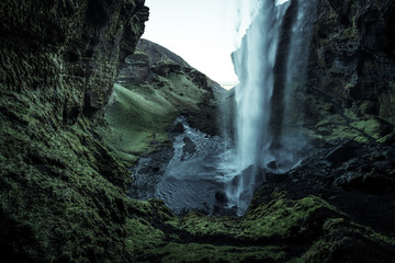 Wild waterfall in a green cave with green moos and wet black sand and stones with a creek flowing through a green grass covered rocky hilly valley surrounded by hills in Iceland